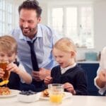 Children Wearing School Uniform In Kitchen Eating Breakfast Waffles As Parents Get Ready For Work