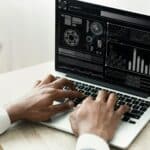 Black Businessman Working On Laptop With Graphs Sitting In Office